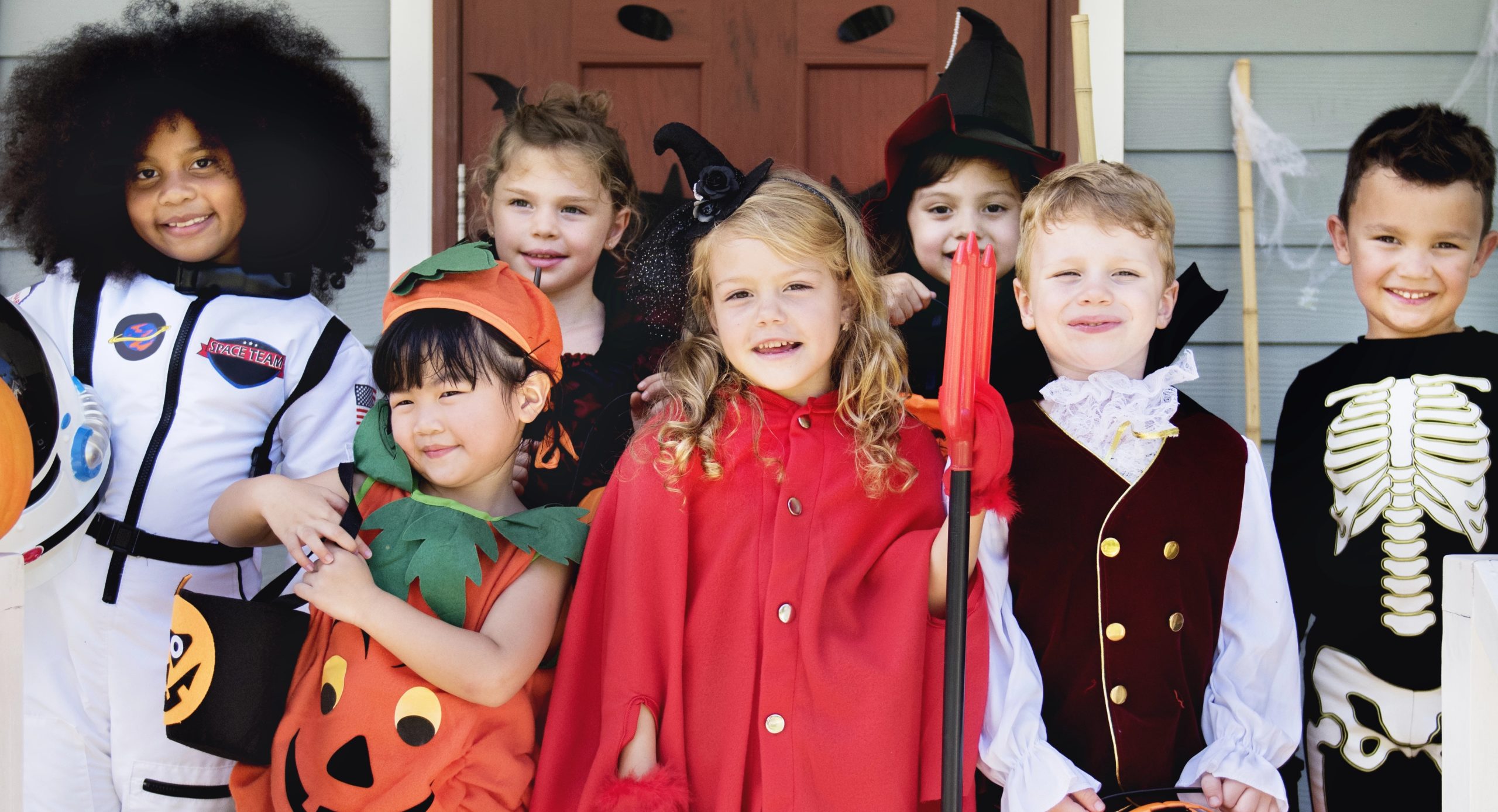 A group of children dressed in friendly Halloween costumes, including a pumpkin, astronaut, witch, and skeleton, smiling together on a front porch.